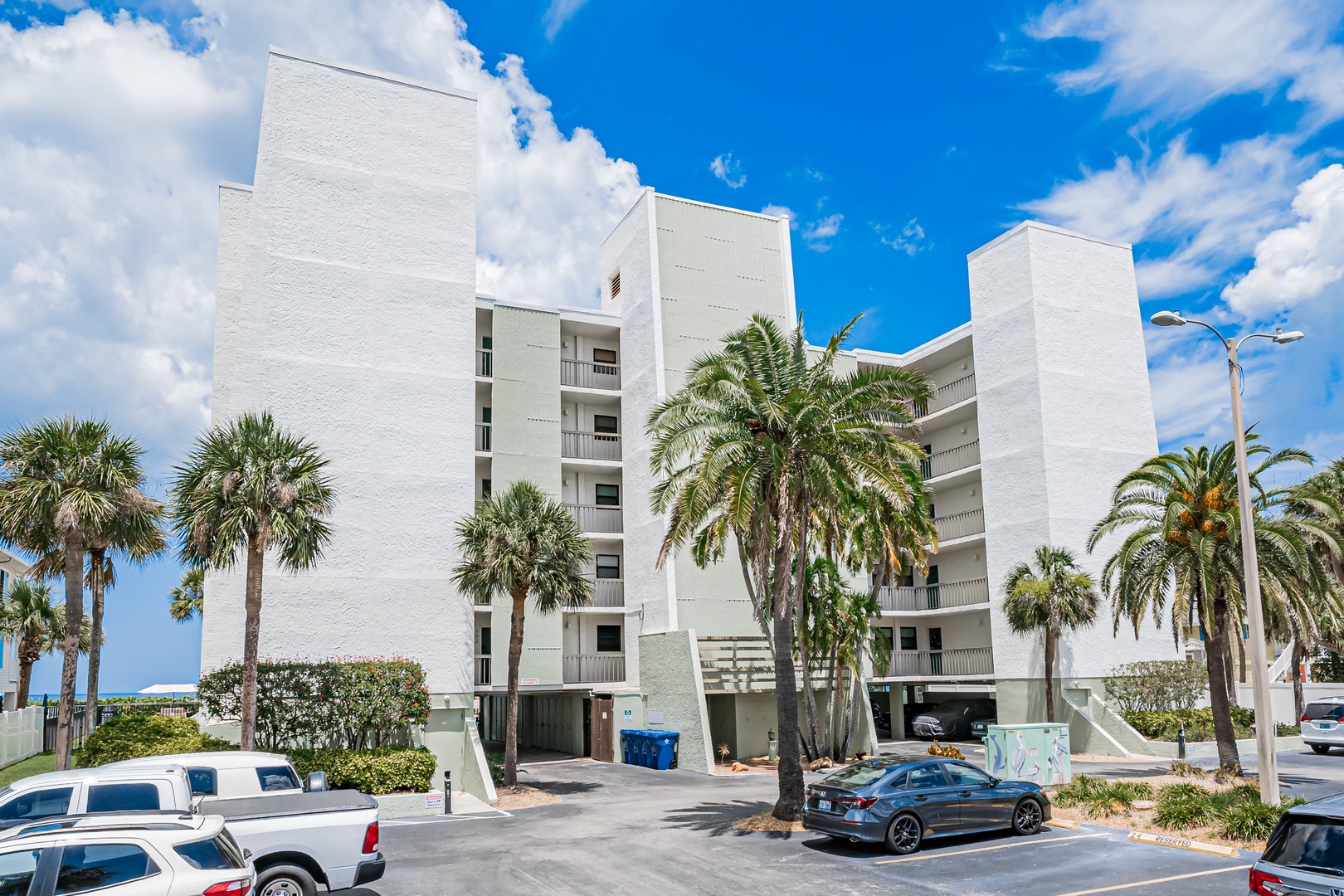 A modern white multi-story building featuring private balconies and tropical palms, with convenient parking and coastal access nearby.