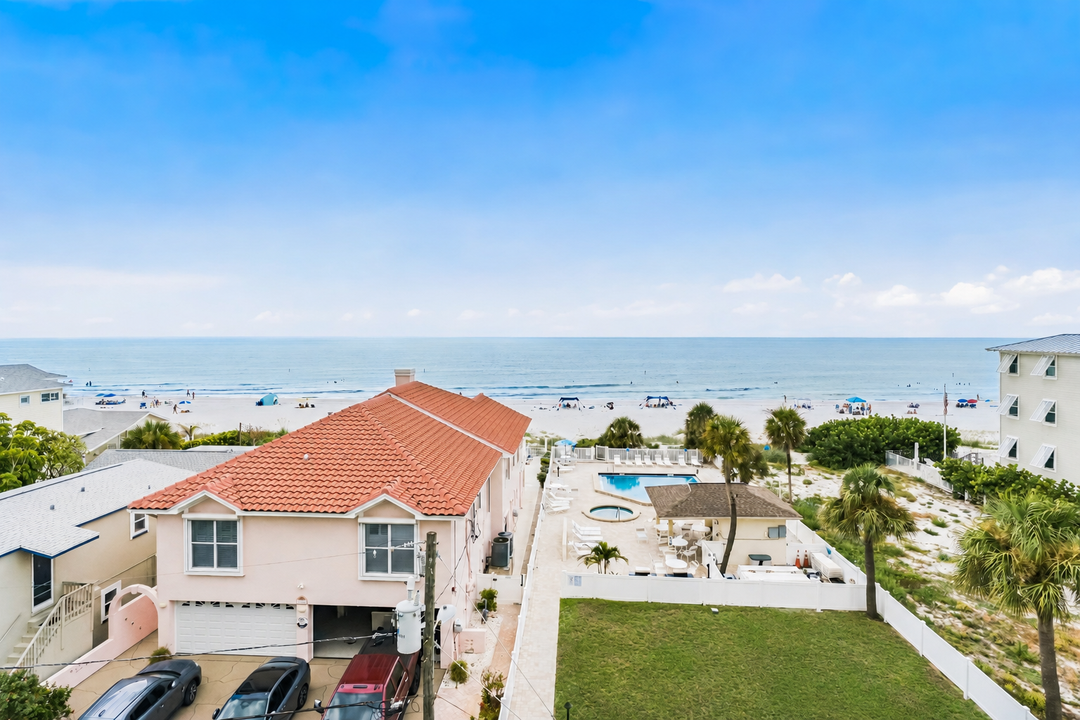 Coastal property set near the beach with sandy shores, gentle waves, and open skies. The property features a pool area surrounded by palms and tropical landscaping.