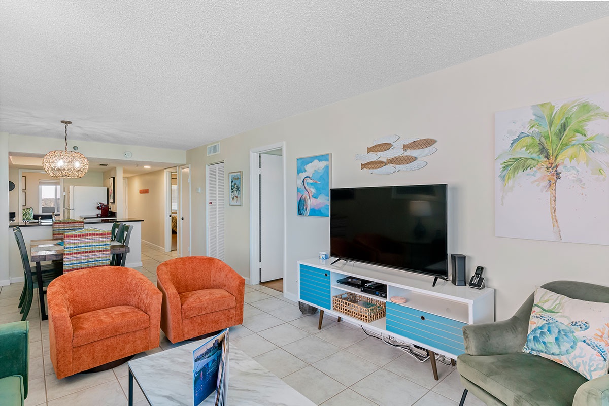 Relax in your coastal living room with vibrant orange chairs, modern TV entertainment center, and tropical artwork creating the perfect beach getaway vibe.