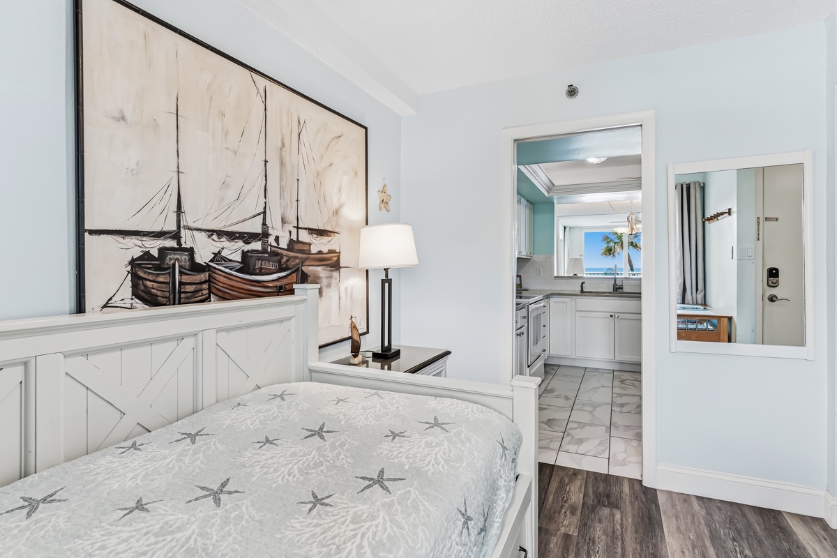 Your coastal retreat awaits with nautical charm and modern comfort. The bedroom flows seamlessly into a bright kitchen where palm trees and blue skies beckon through the window.