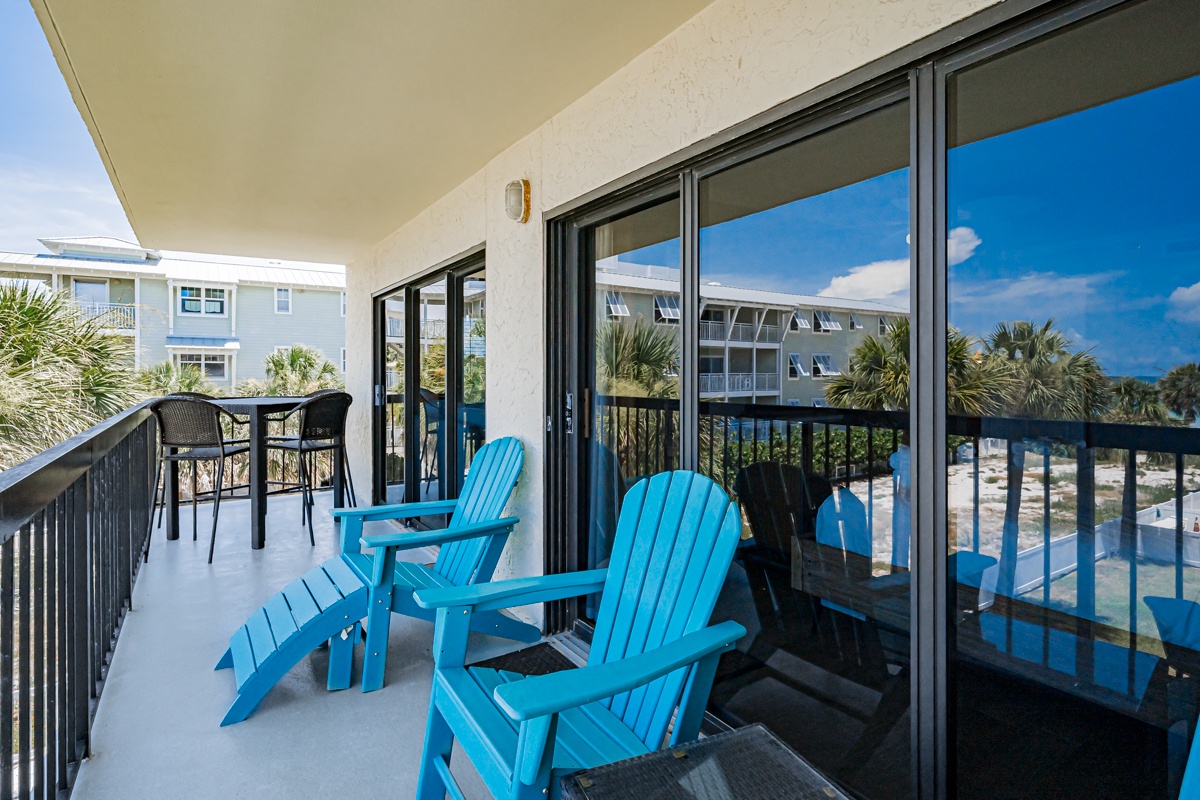 A spacious balcony overlooks tropical palms and neighboring buildings in a coastal setting, offering multiple seating areas for relaxation and outdoor dining.