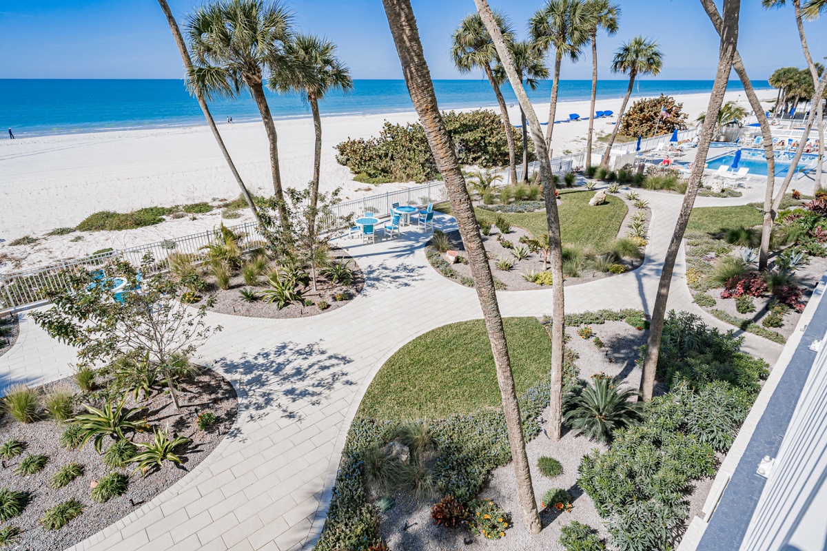 Landscaped grounds wind through towering palms toward a pristine beachfront, where soft sand and turquoise waters meet beneath clear blue skies.