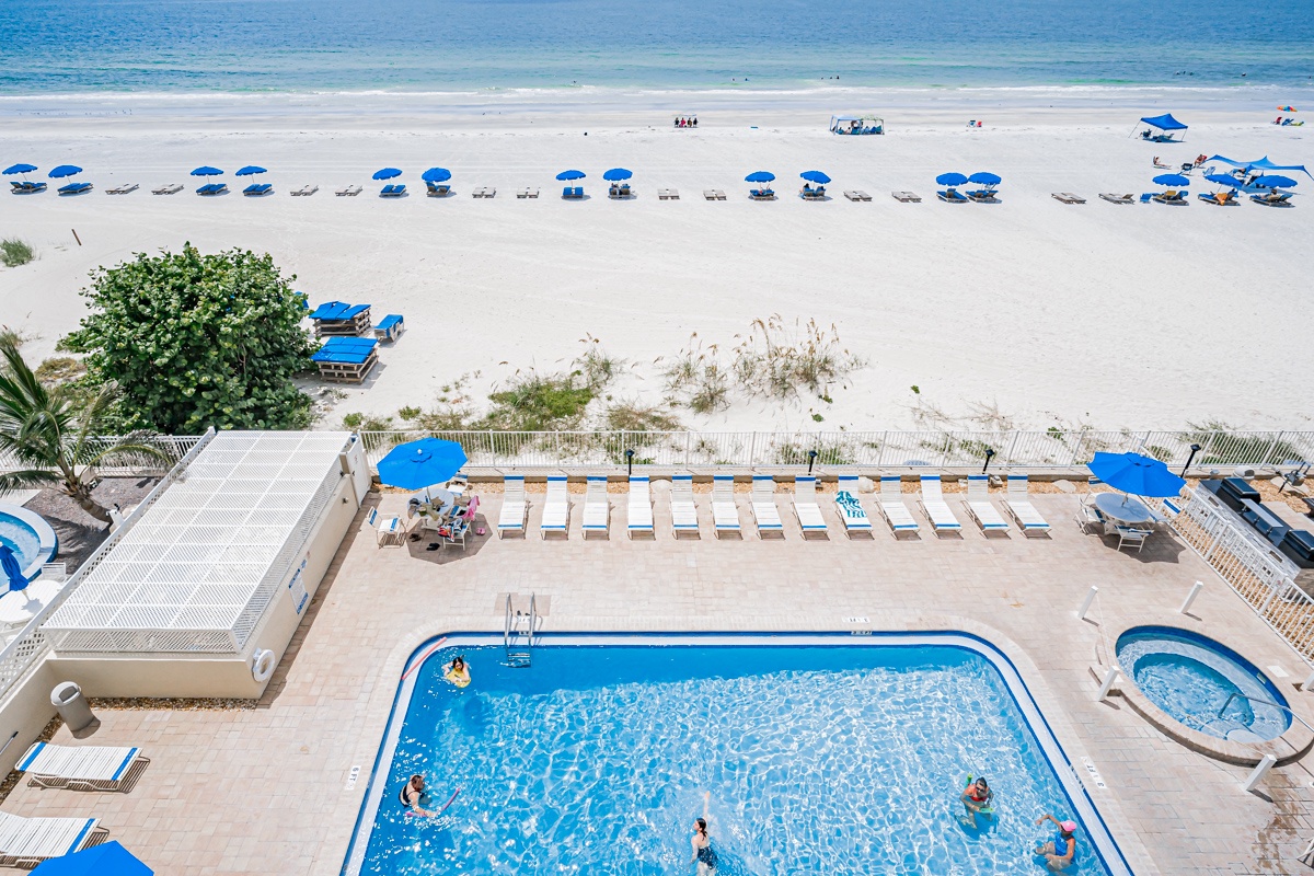 This aerial view captures the beachfront property with its inviting pool area, sun deck with loungers, and direct access to the sandy beach, where rows of umbrellas and chairs await beachgoers along the sparkling shoreline.