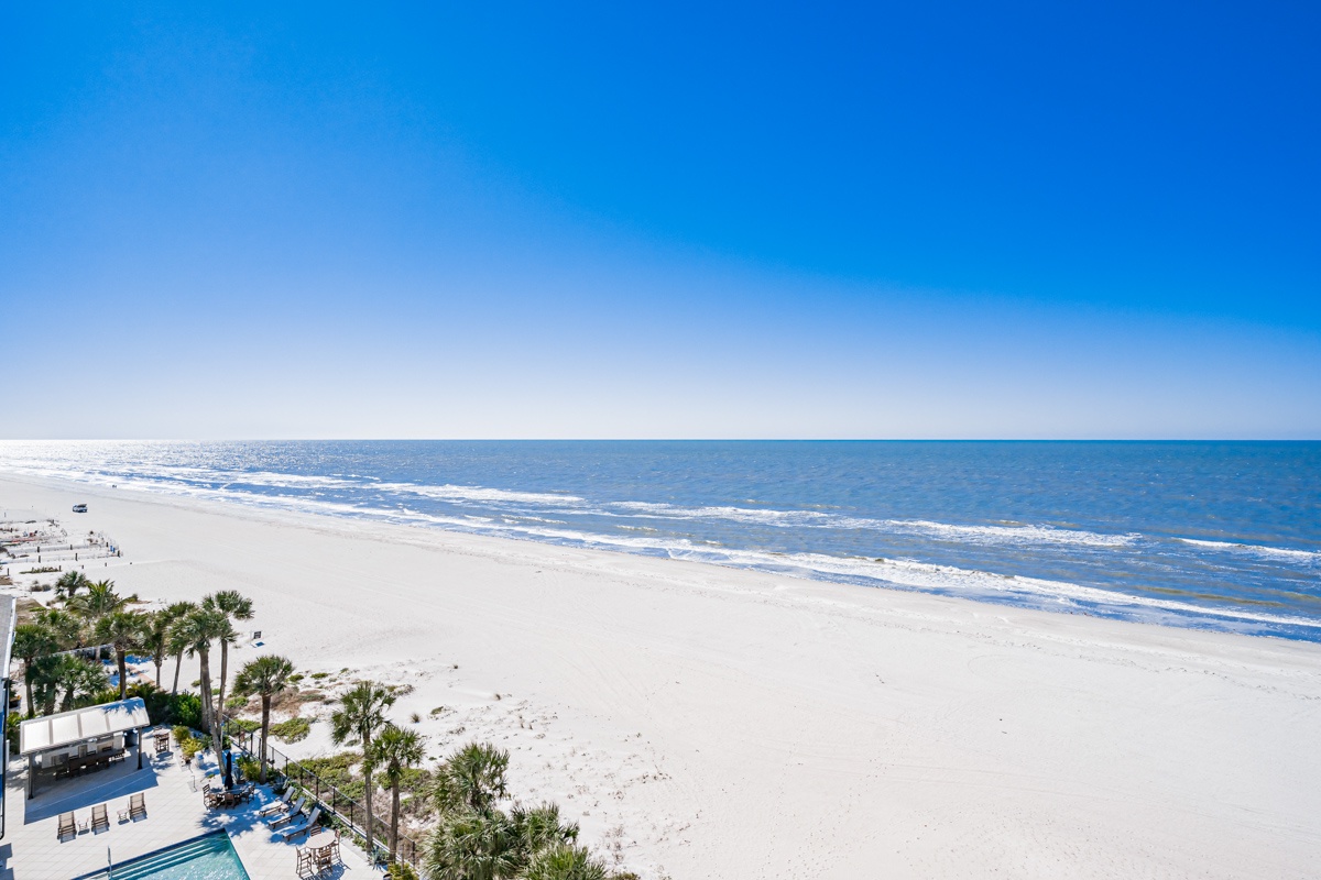 Pristine white sand beach stretches endlessly along crystal-clear waters under brilliant blue skies, with tropical palm trees lining the shore.