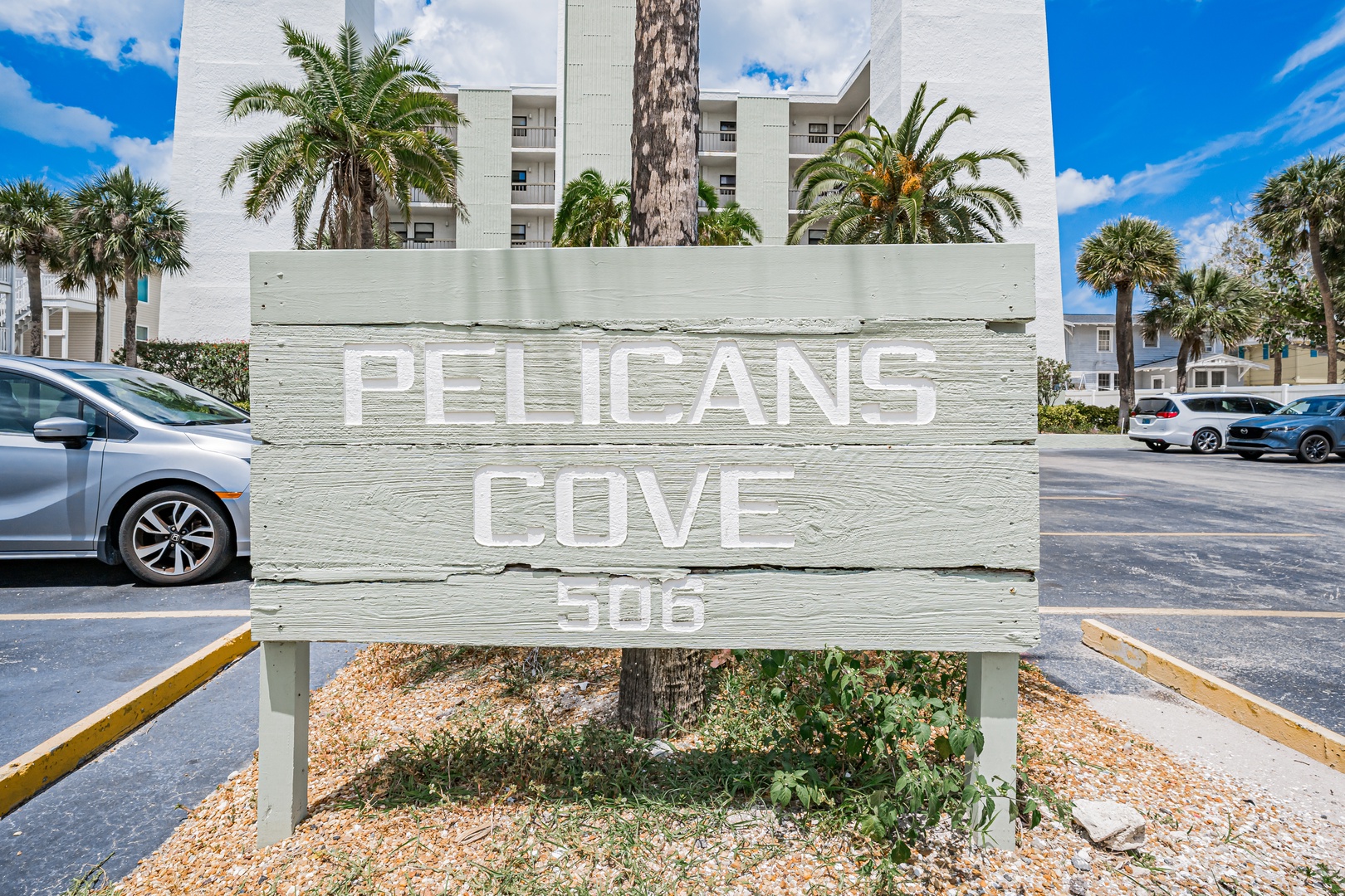 Welcoming property sign for Pelicans Cove 505 set against palm trees and the main building in a serene tropical location.