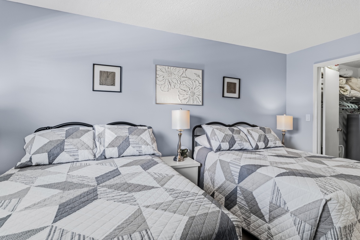 Your serene bedroom awaits with two plush Double beds dressed in elegant geometric quilts and soft pillows. Bedside lamps cast a warm glow as you settle in for a restful night.