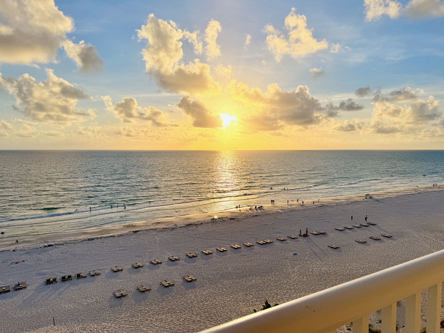 A stunning beachfront location where the sun sets over calm turquoise waters and soft white sand. Rows of lounge chairs line the shore, perfect for soaking in the coastal beauty.