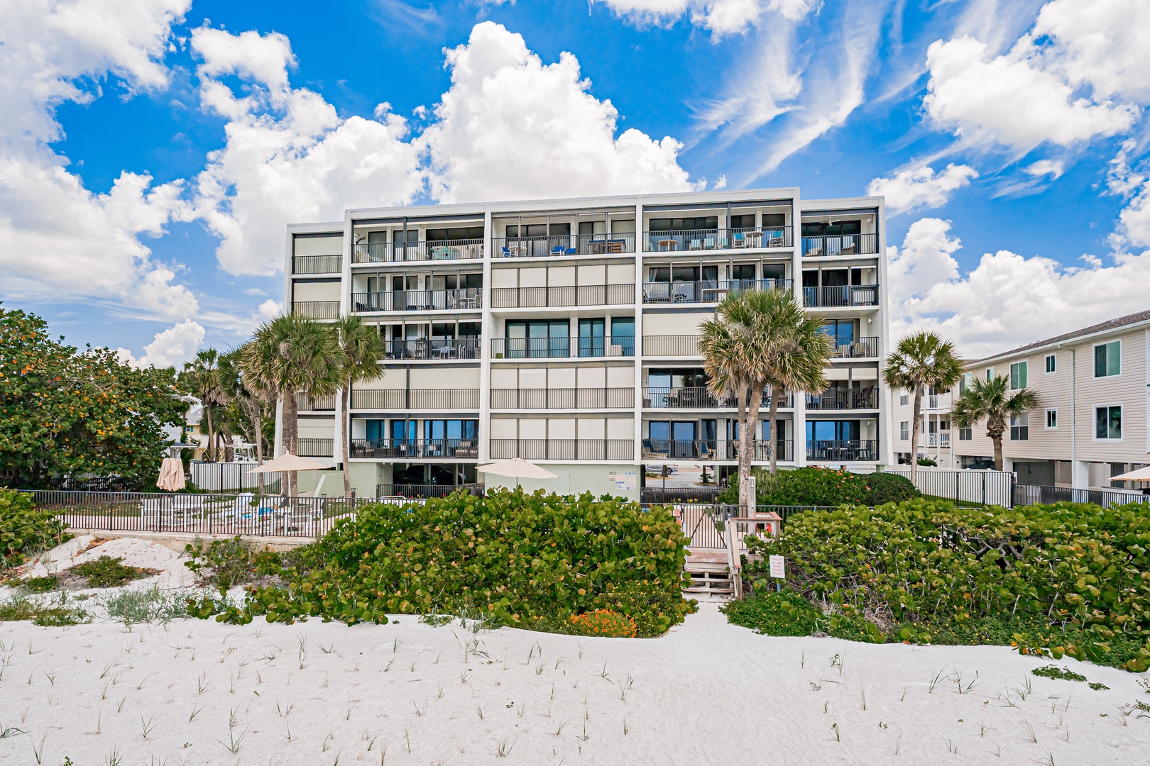 Beachfront property with direct white sand access, tropical landscaping, and multiple private balconies overlooking the ocean under a bright blue sky.