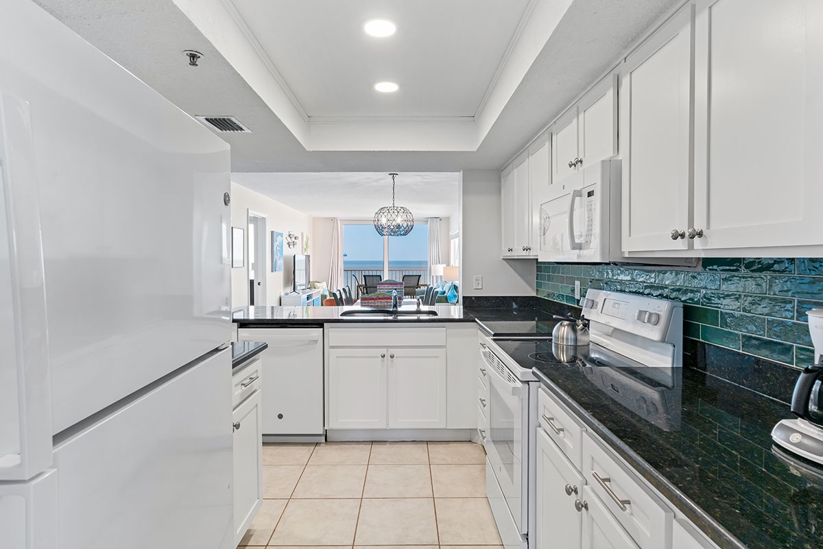 Your fully equipped kitchen features stunning emerald tile backsplash, granite counters, and ocean glimpses beyond the living area.