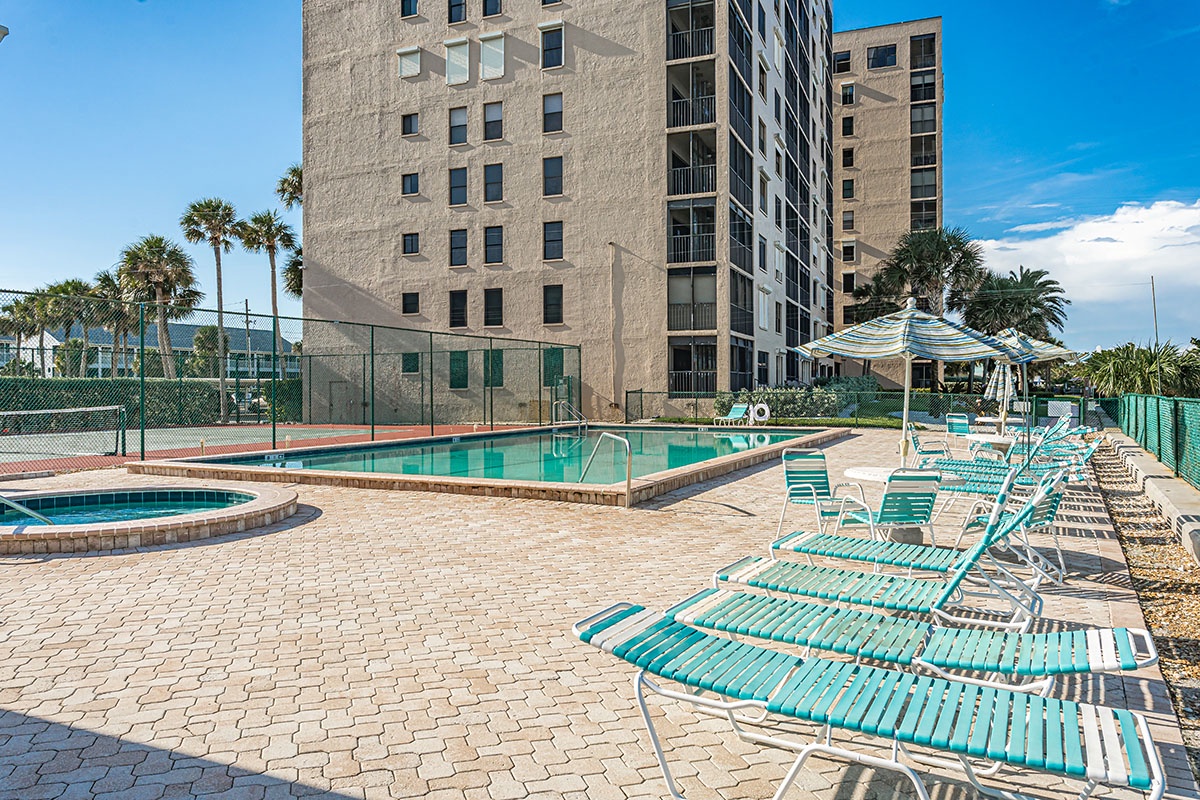 Modern resort-style pool deck with comfortable loungers and tennis court, surrounded by palm trees under sunny skies.