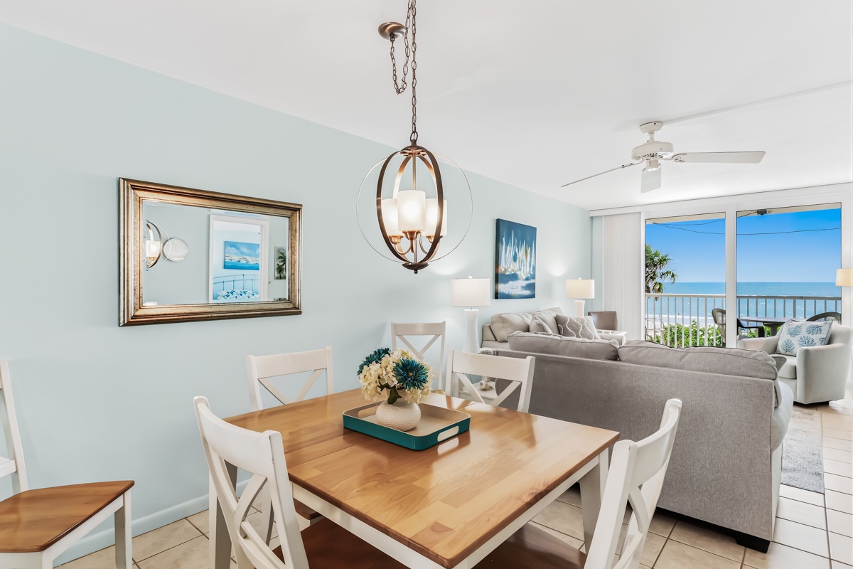 Your dining area flows seamlessly into the living space, where soft coastal blues meet warm wood tones and a stunning ocean view awaits through the balcony doors.