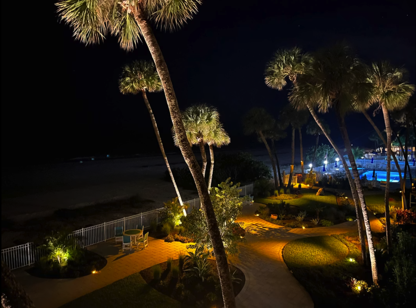 Illuminated palm trees frame a beautifully lit pathway leading to the waterfront, with the pool and surrounding grounds glowing under the evening sky.
