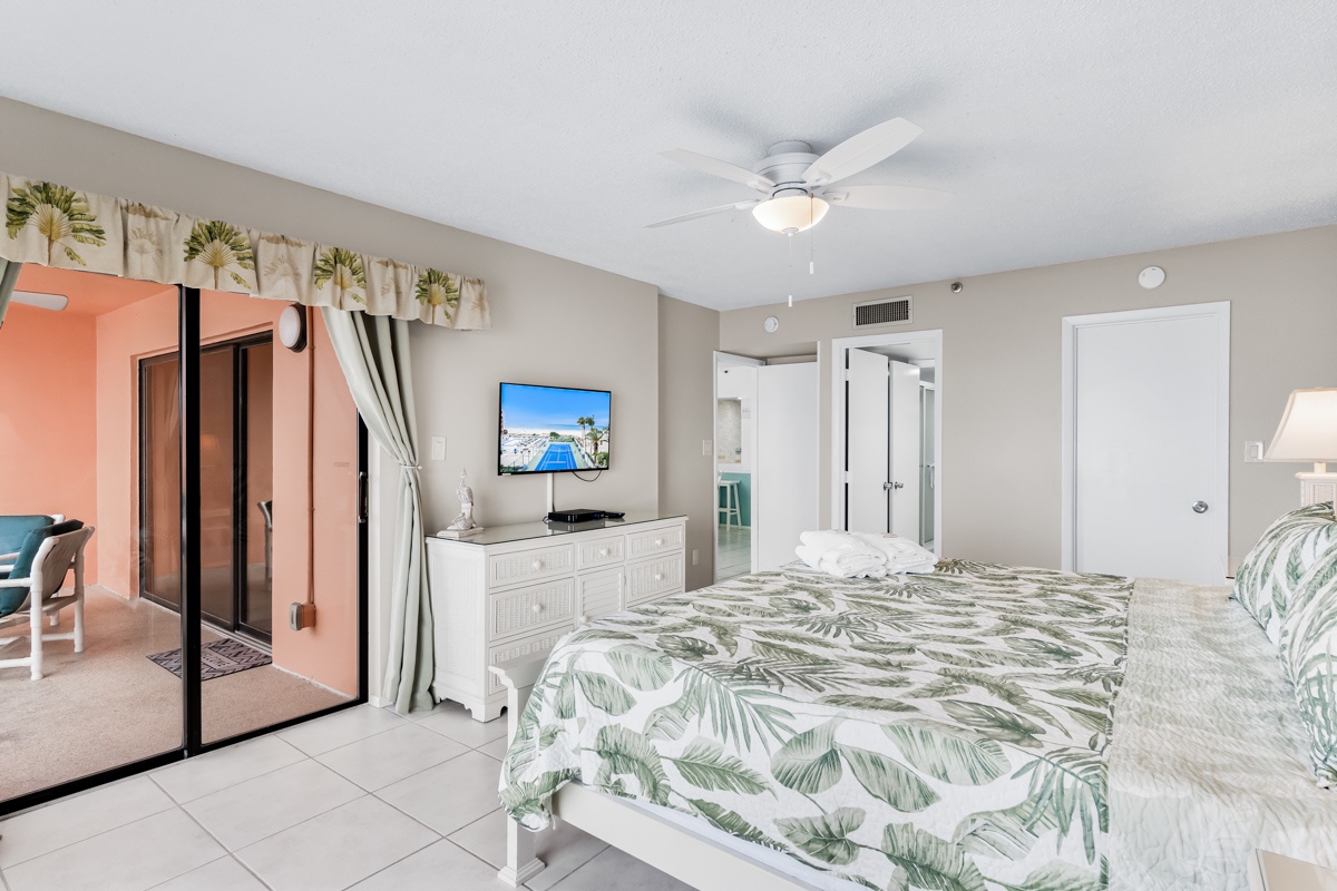Your tropical bedroom retreat features palm-themed linens, sliding balcony doors, and modern TV entertainment for ultimate beachside relaxation.