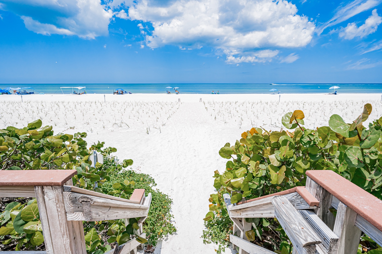 A scenic beachfront area with white sand and turquoise waters, framed by coastal vegetation. The property offers direct access to a pristine shoreline with calm seas stretching to the horizon.