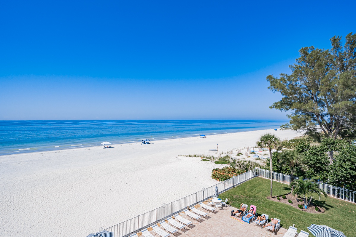 A pristine beachfront stretches along calm, azure waters under bright skies, offering direct access to a wide sandy shore perfect for lounging and enjoying the coastal scenery.
