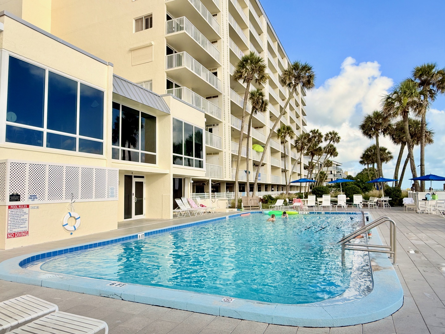 The property features an outdoor swimming pool surrounded by palm trees and ample lounge seating, with direct beach access just beyond the pool deck.