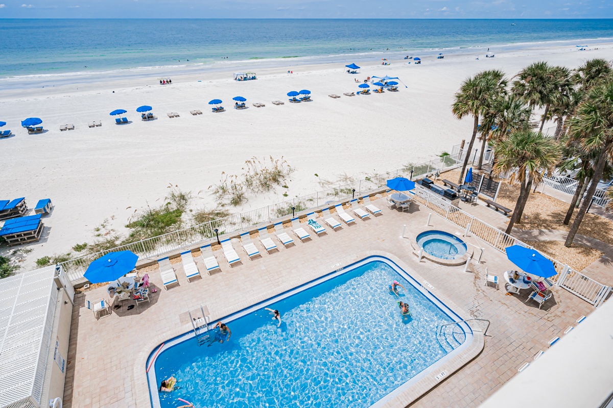 Aerial view of beachfront property pool area and pristine white sand beach with rows of blue umbrellas along the shoreline.