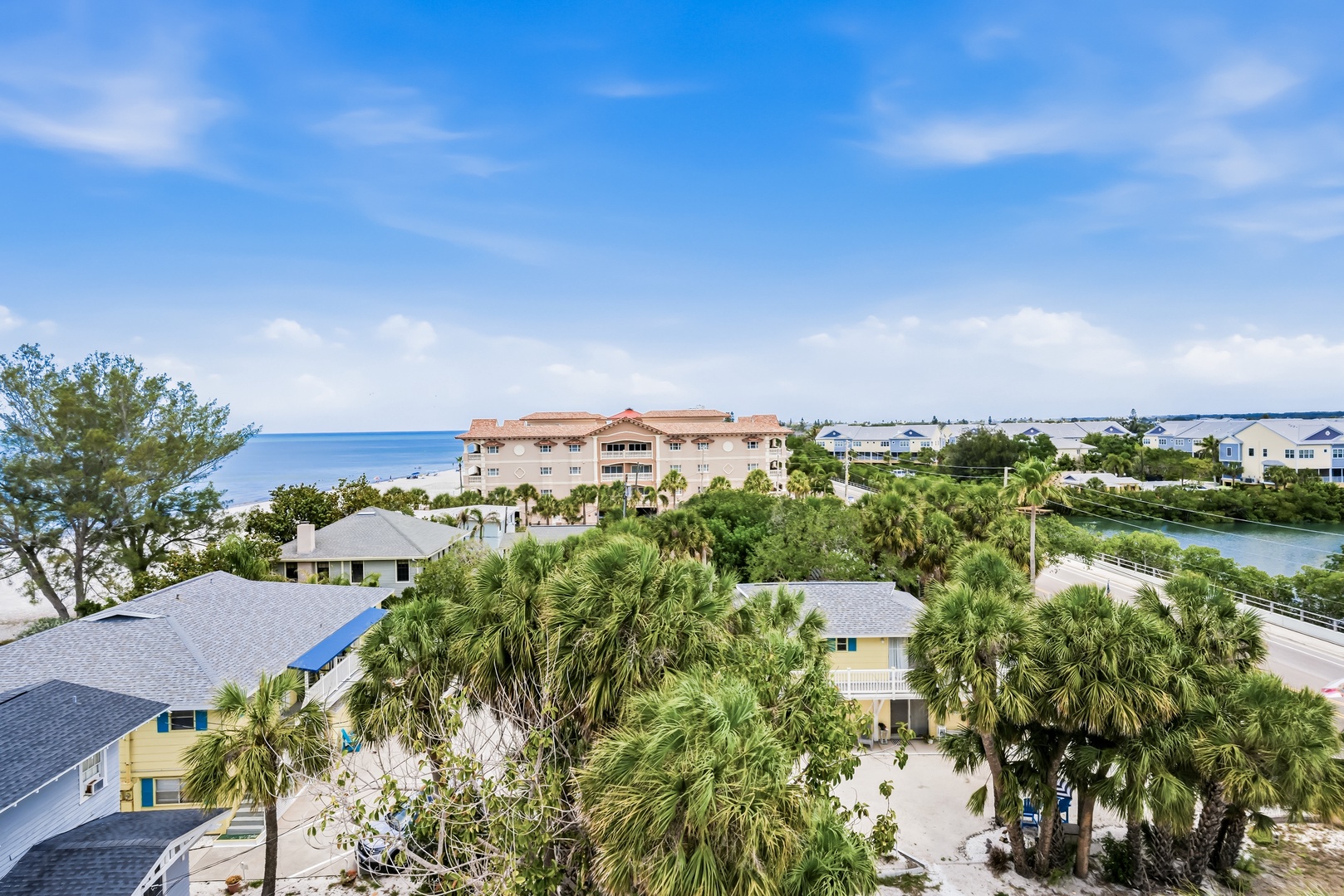Aerial view captures the property's coastal setting, with tropical palms framing the buildings and sparkling blue water stretching to the horizon beyond nearby residential areas.