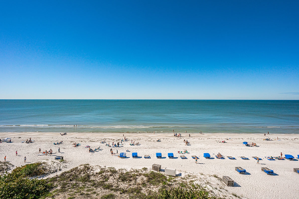 Pristine white sand beach stretches along turquoise waters under endless blue skies, with beachgoers enjoying the coastal paradise.