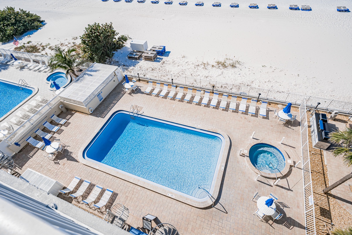 Beachfront property features sparkling pool area with hot tub, sun deck with lounge chairs, and direct white sand beach access with blue umbrella service.