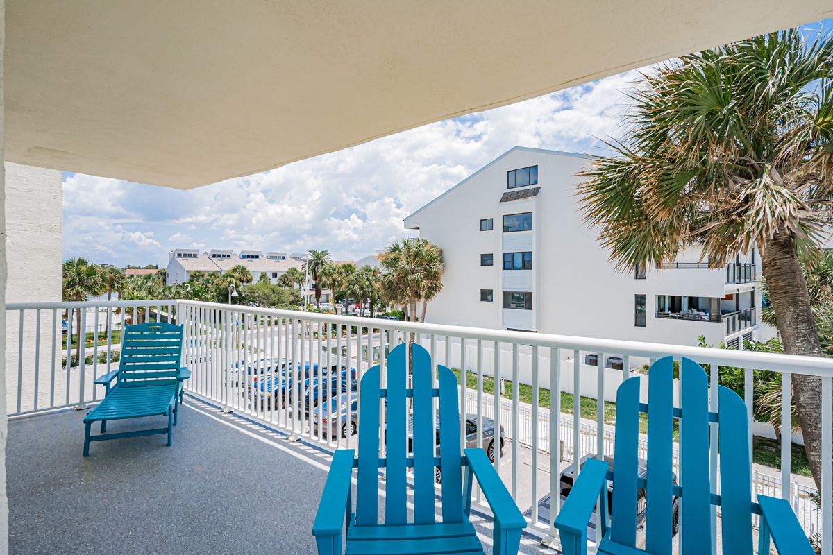 The extra spacious corner wrap balcony at this beachfront property features colorful Adirondack-style chairs and a lounge chair, framed by swaying palm trees and nearby buildings.