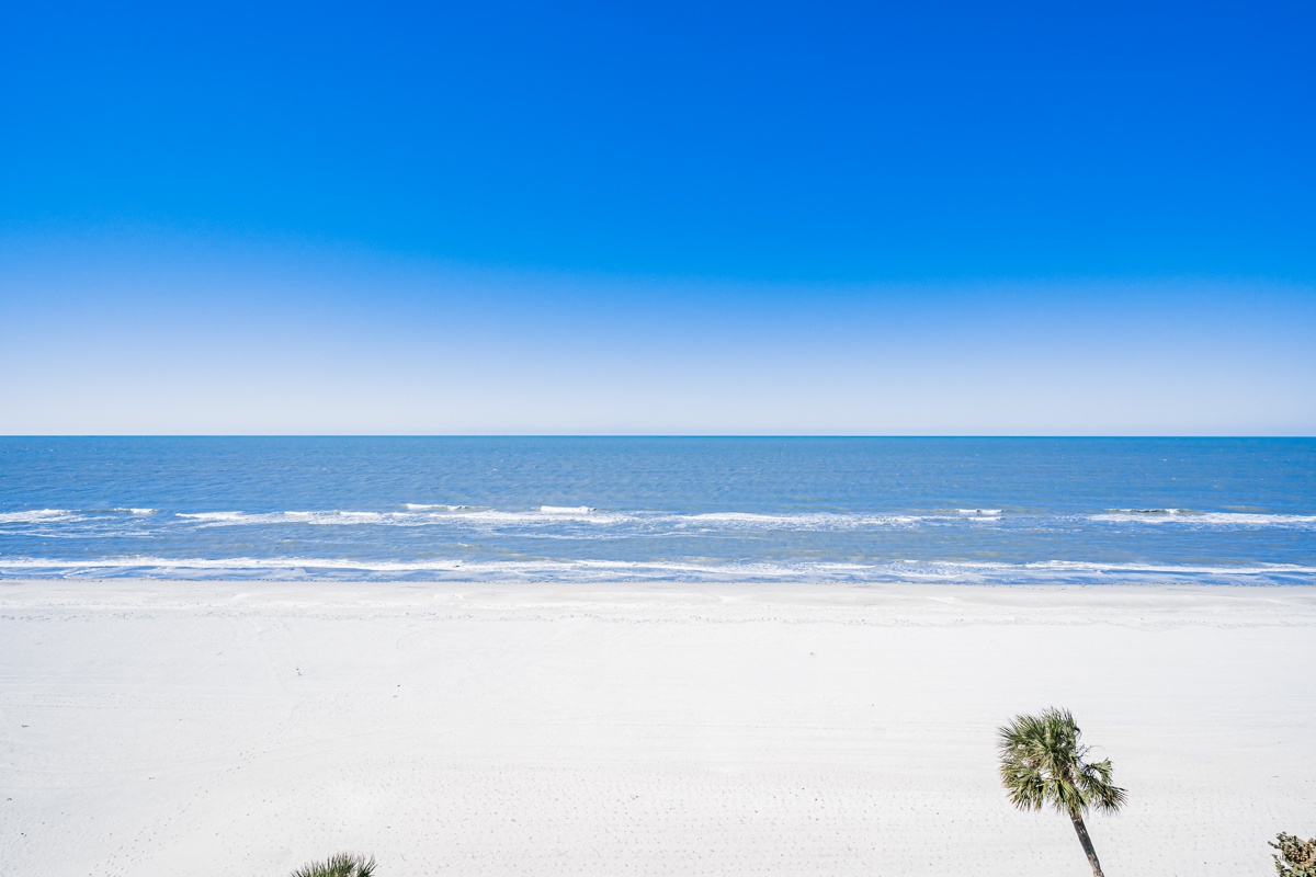 Pristine white sand beach stretches endlessly under brilliant blue skies, with gentle waves rolling onto shore and tropical palms swaying nearby.