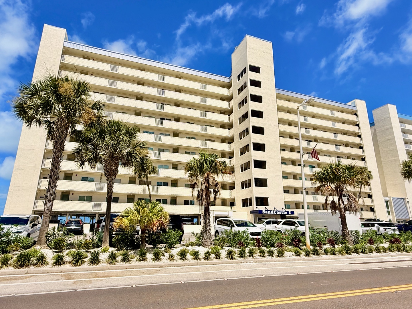 A mid-rise beachfront building with palm trees and tropical landscaping, set against a backdrop of blue skies and fluffy clouds, marking the entrance to your coastal getaway.