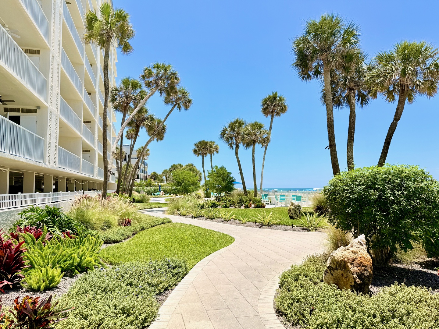 Winding pathways lead you through lush tropical landscaping toward the sparkling turquoise waters, where swaying palms and coastal breezes set the scene for your perfect beach escape.