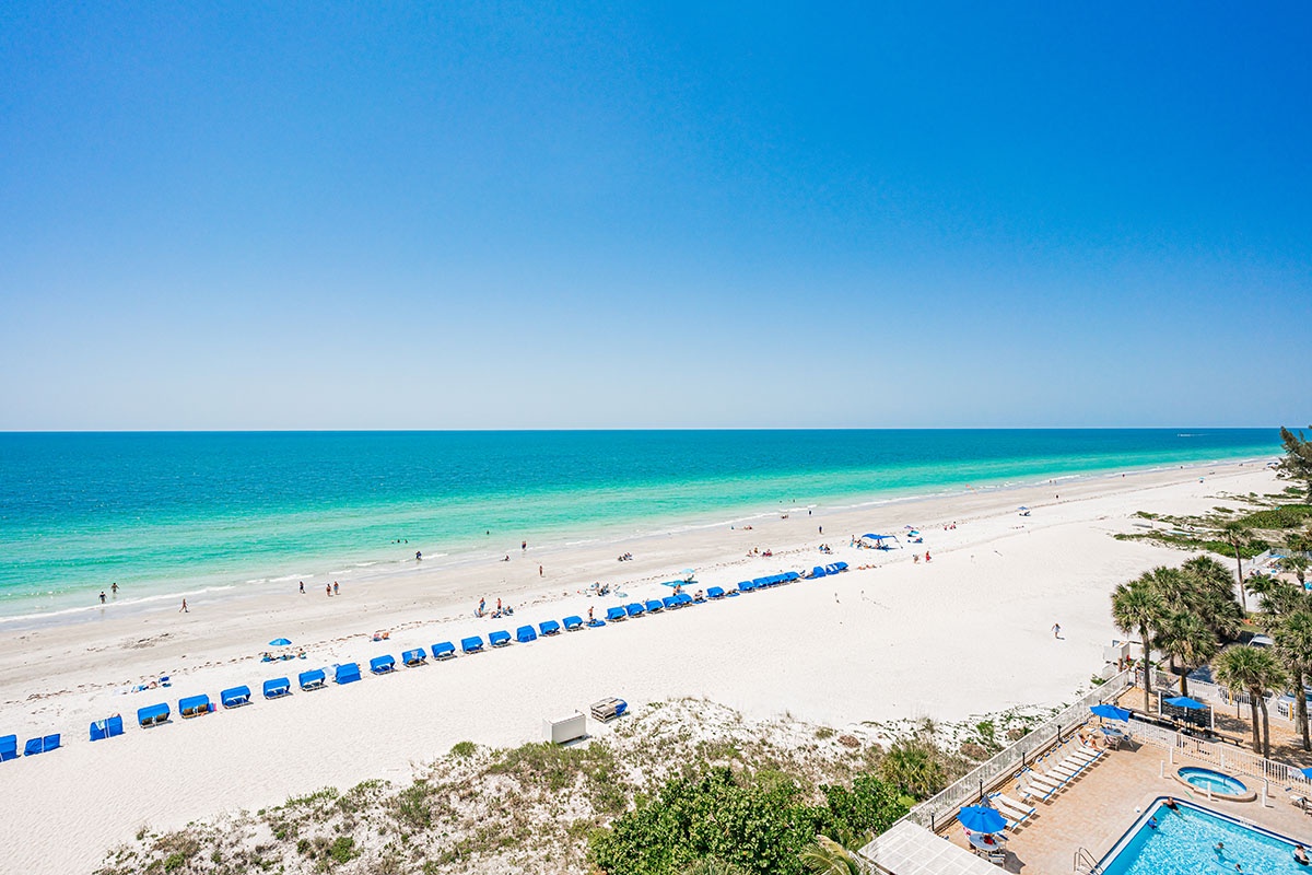 Pristine white sand beach stretches along turquoise waters with organized blue umbrellas and palm trees dotting the coastline.