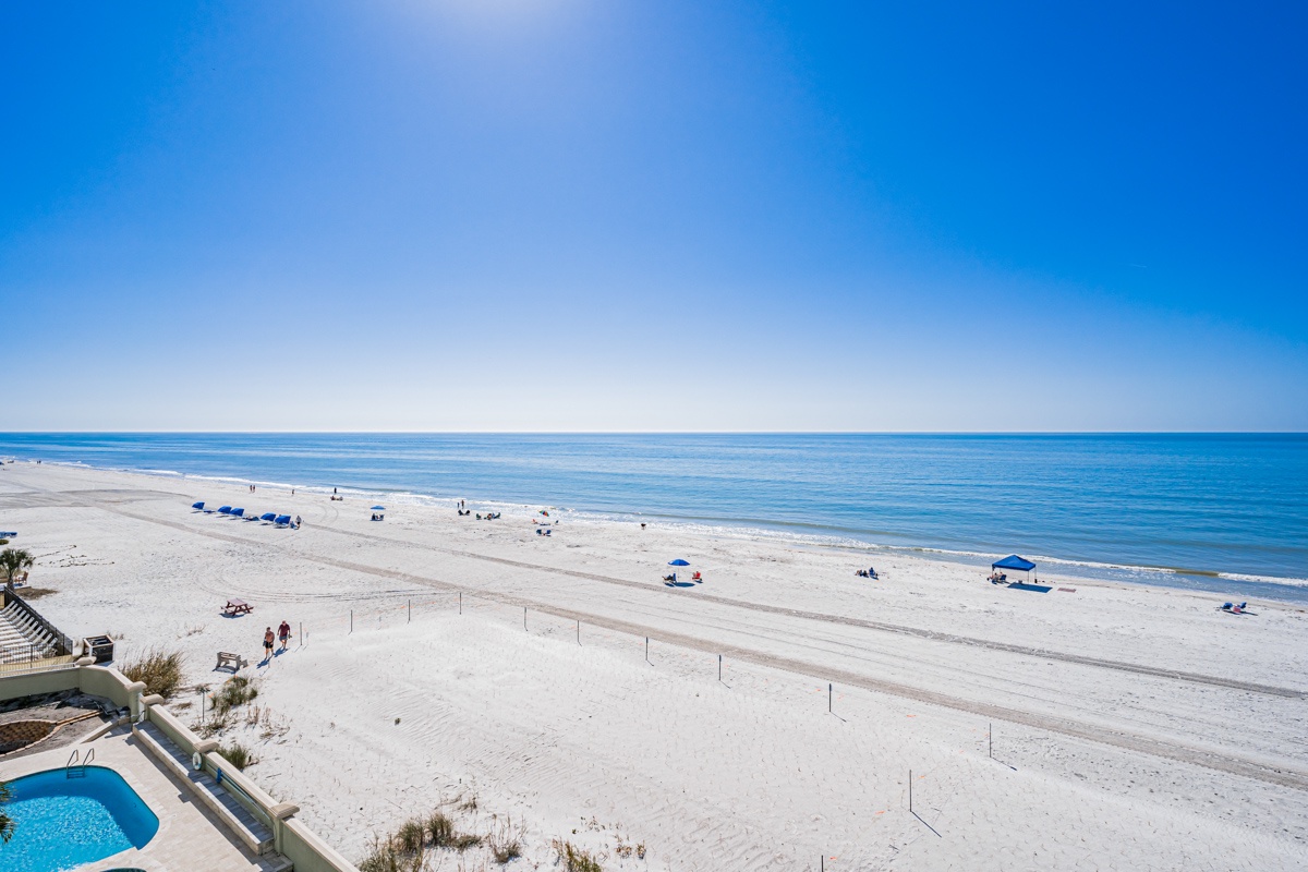 Looking southward. Pristine white sand beach stretching along crystal-clear turquoise waters under brilliant blue skies, with beachgoers enjoying the coastal paradise.