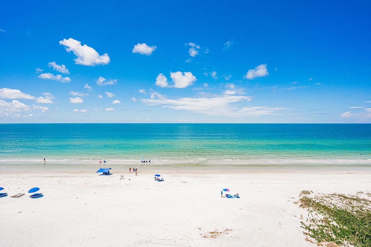 Pristine white sand beach stretches endlessly along turquoise waters under brilliant blue skies dotted with fluffy clouds.
