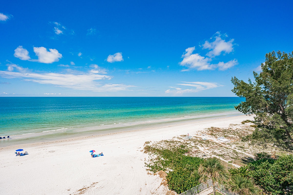Pristine white sand beach stretches along turquoise waters under brilliant blue skies with scattered beachgoers enjoying the coastal paradise.