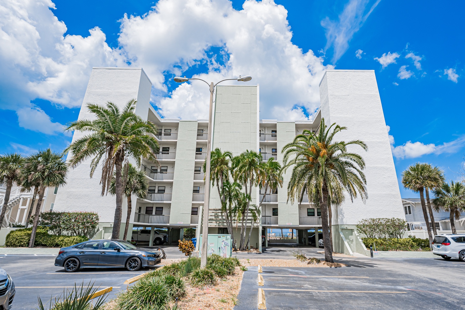 Tropical palm trees line the property entrance, while covered parking below offers convenient access to the multi-story beachfront building under bright Florida skies.