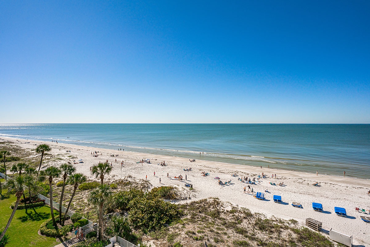 Pristine white sand beach stretches along turquoise waters under brilliant blue skies, with palm trees and beachgoers enjoying the coastal paradise.