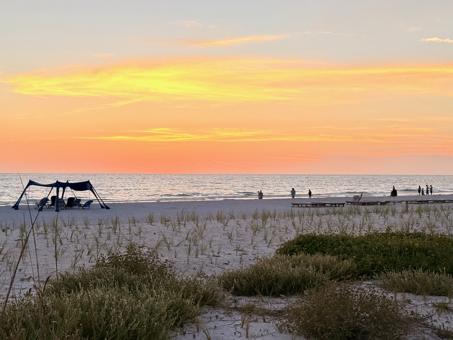Golden hour paints the sky in stunning coral and amber hues as the day winds down along the sandy shore, with the calm sea stretching to the horizon—a tranquil scene perfect for evening walks.
