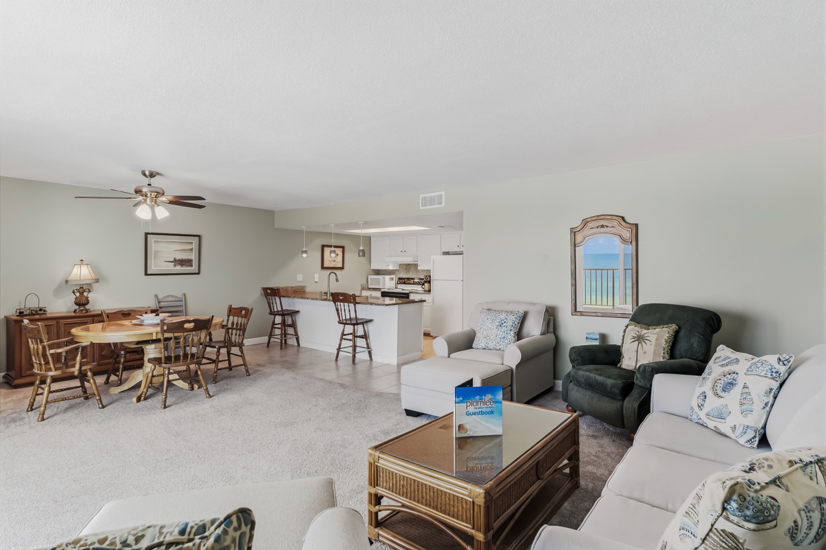 Relax in this spacious open-concept living area featuring soft carpeting, comfortable seating, and a bright kitchen perfect for your beach getaway. Notice the stunning sea view through the window, inviting you to unwind with coastal tranquility.