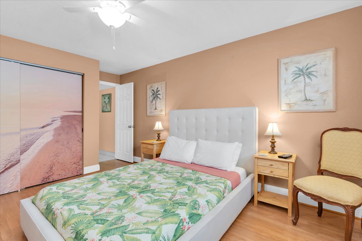 Your coastal retreat features a plush upholstered Queen bed dressed in tropical linens, warm peach walls adorned with palm artwork, and soft bedside lighting for ultimate relaxation.