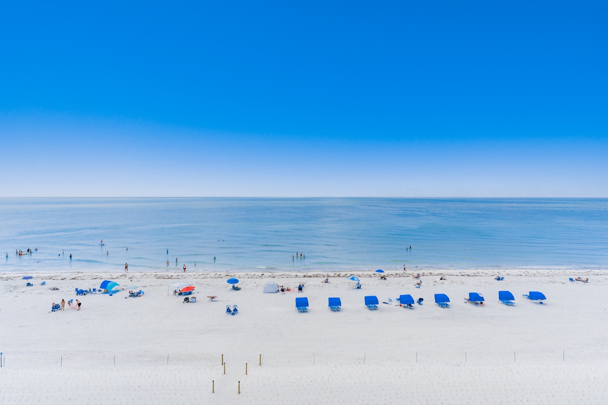 Pristine white sand beach stretches endlessly under brilliant blue skies, with scattered umbrellas and beachgoers enjoying the Gulf waters.