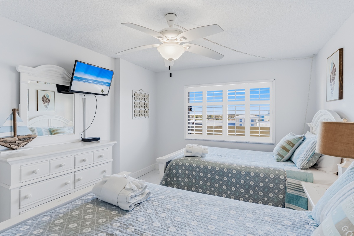 Unwind in your coastal bedroom with queen beds, ceiling fan, and bright windows overlooking the area.