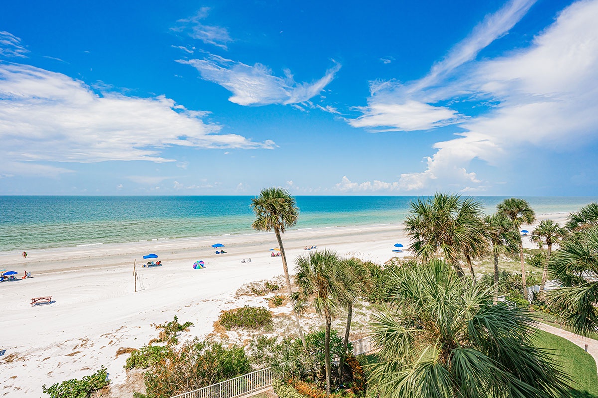 Pristine white sand beach stretches along turquoise waters, framed by swaying palms and brilliant blue skies with scattered beachgoers enjoying the tropical paradise.