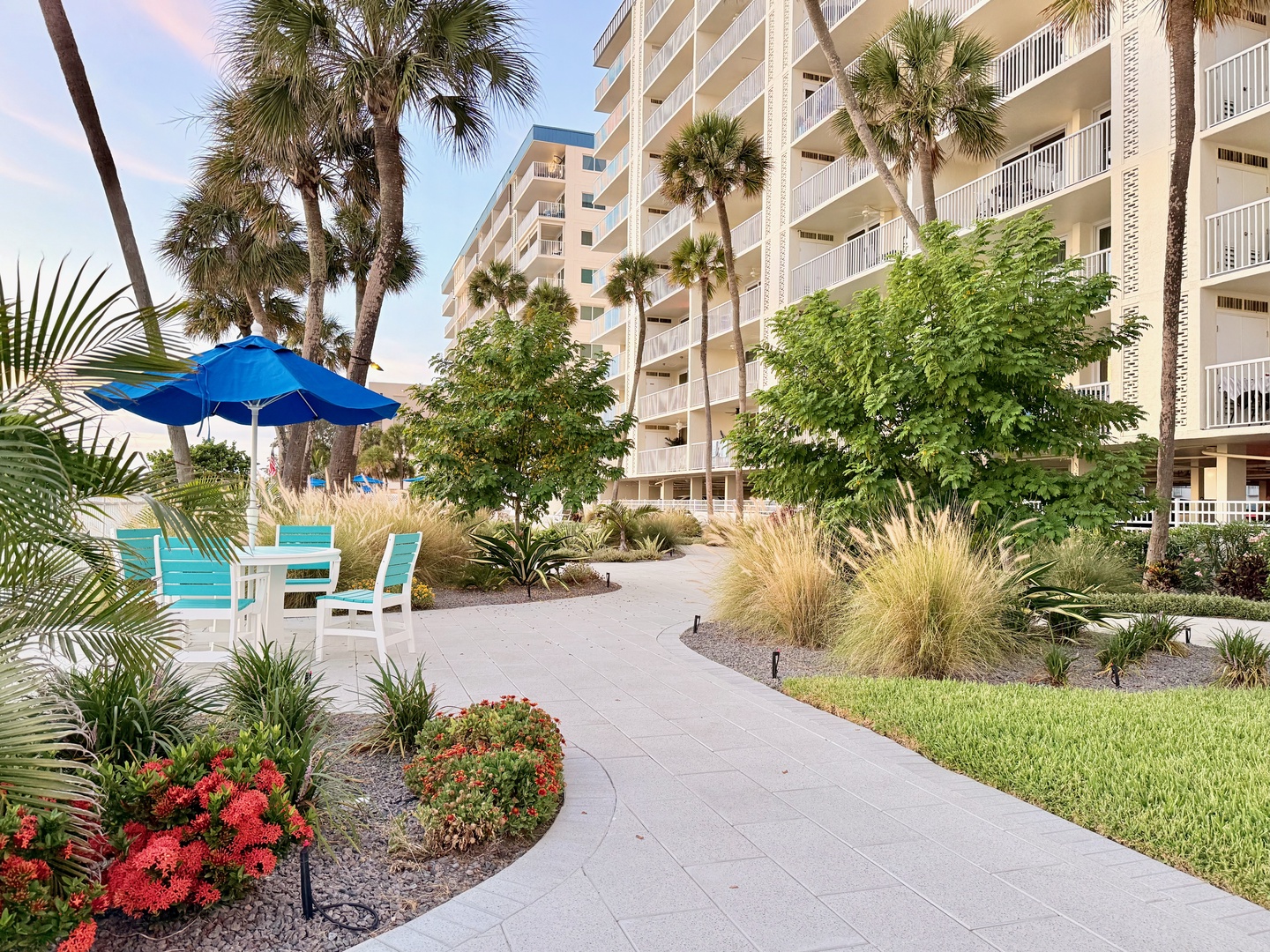 The property sits nestled among palm trees and lush landscaping, with winding pathways leading past vibrant tropical gardens and inviting outdoor seating areas under the Florida sun.