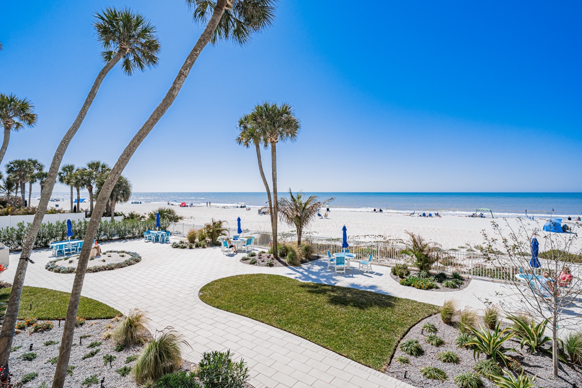 Tropical palm trees frame pristine white sand beach with blue umbrellas and landscaped grounds at this beachfront property.