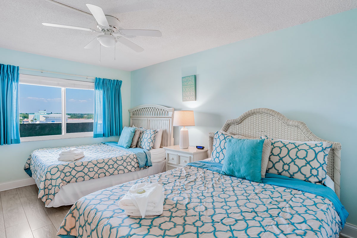 Your coastal-themed bedroom features two comfortable beds with turquoise accents, ceiling fan, and bright windows for a refreshing seaside retreat.