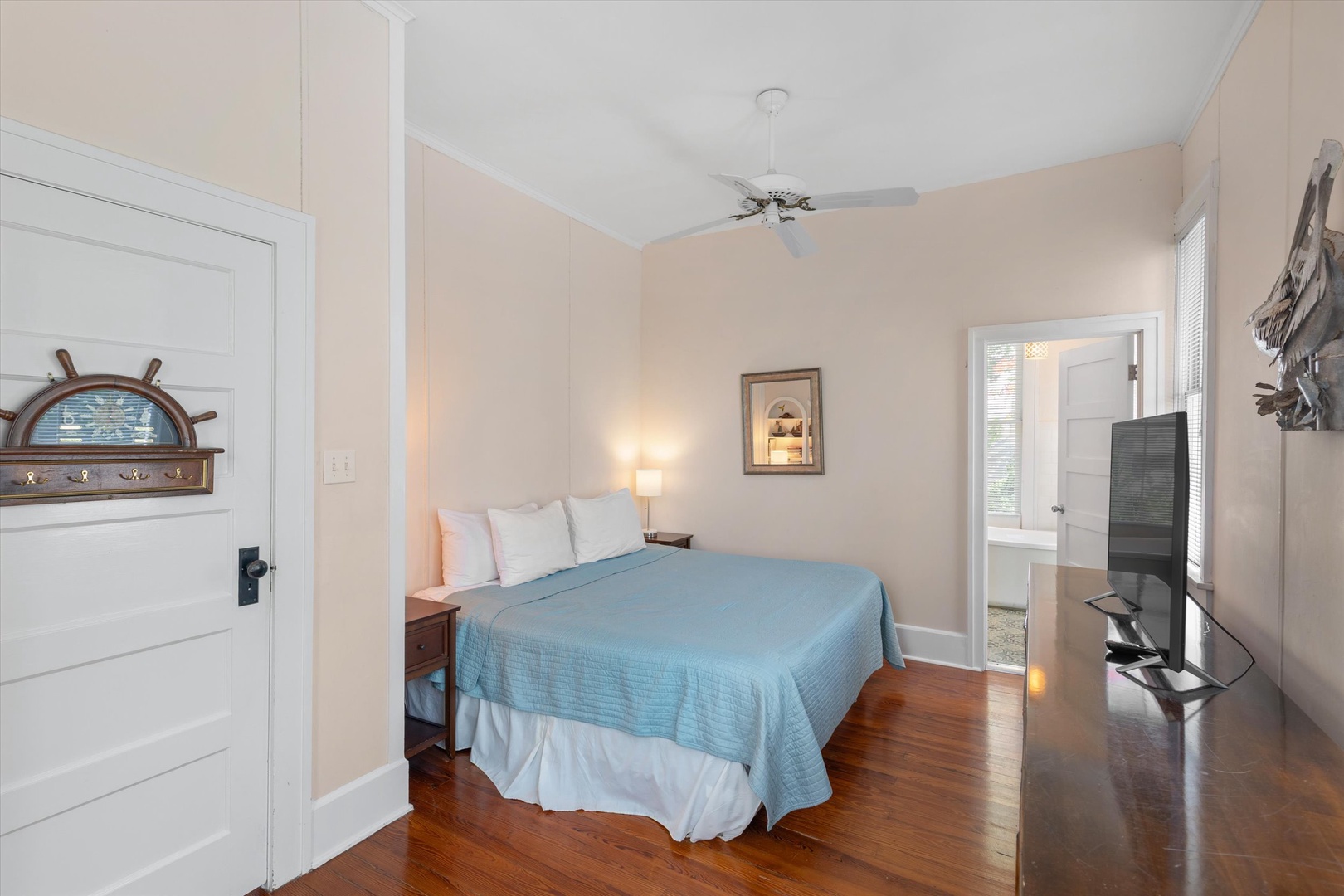 Relax in this serene coastal bedroom with calming beachy tones, soft lighting, and nautical charm. A comfortable bed awaits your restful nights, while the ceiling fan keeps you cool. Natural hardwood floors and decorative touches create a welcoming retrea