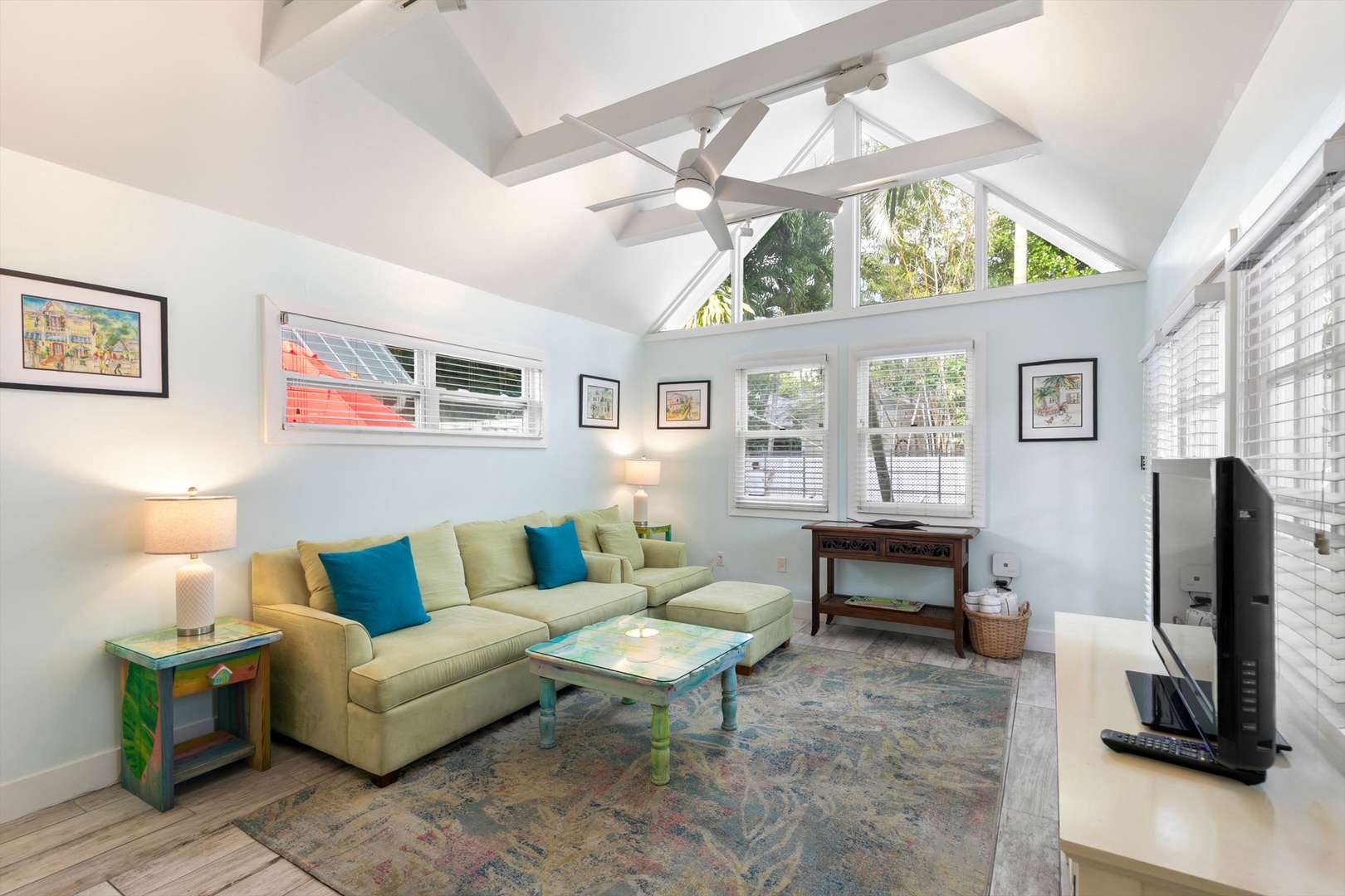 Bright, airy living room with soaring vaulted ceilings and abundant windows. The soft green sofa invites relaxation beneath ceiling fans, while natural light frames lush garden views.