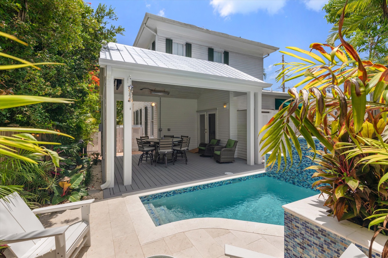 A white two-story home with metal roof stands surrounded by lush tropical gardens, featuring a covered patio and sparkling pool for relaxation and outdoor dining.