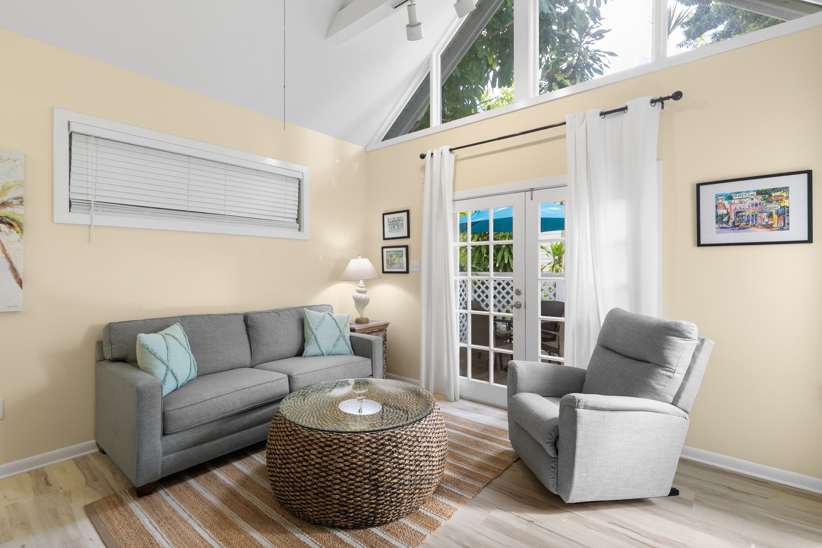 Relax in your sunlit living room where soft gray furnishings and soaring ceilings create an airy, welcoming retreat. Glass doors open to glimpses of tropical greenery beyond, inviting you to unwind in coastal comfort.