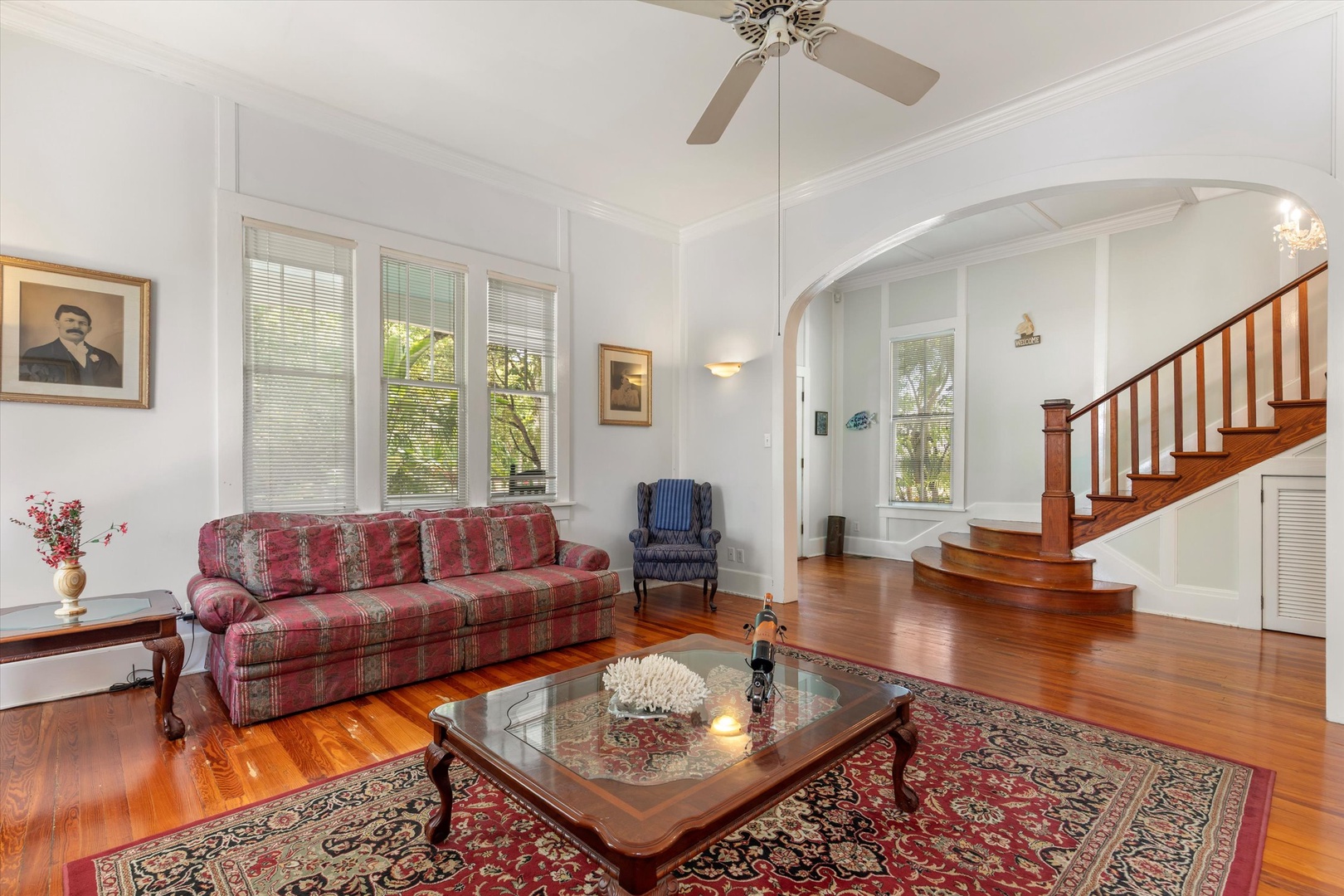 Relax in this bright, spacious living room featuring stunning hardwood floors, soaring ceilings with elegant crown molding, and a beautiful wooden staircase. Large windows flood the space with natural light, creating a warm and welcoming atmosphere.