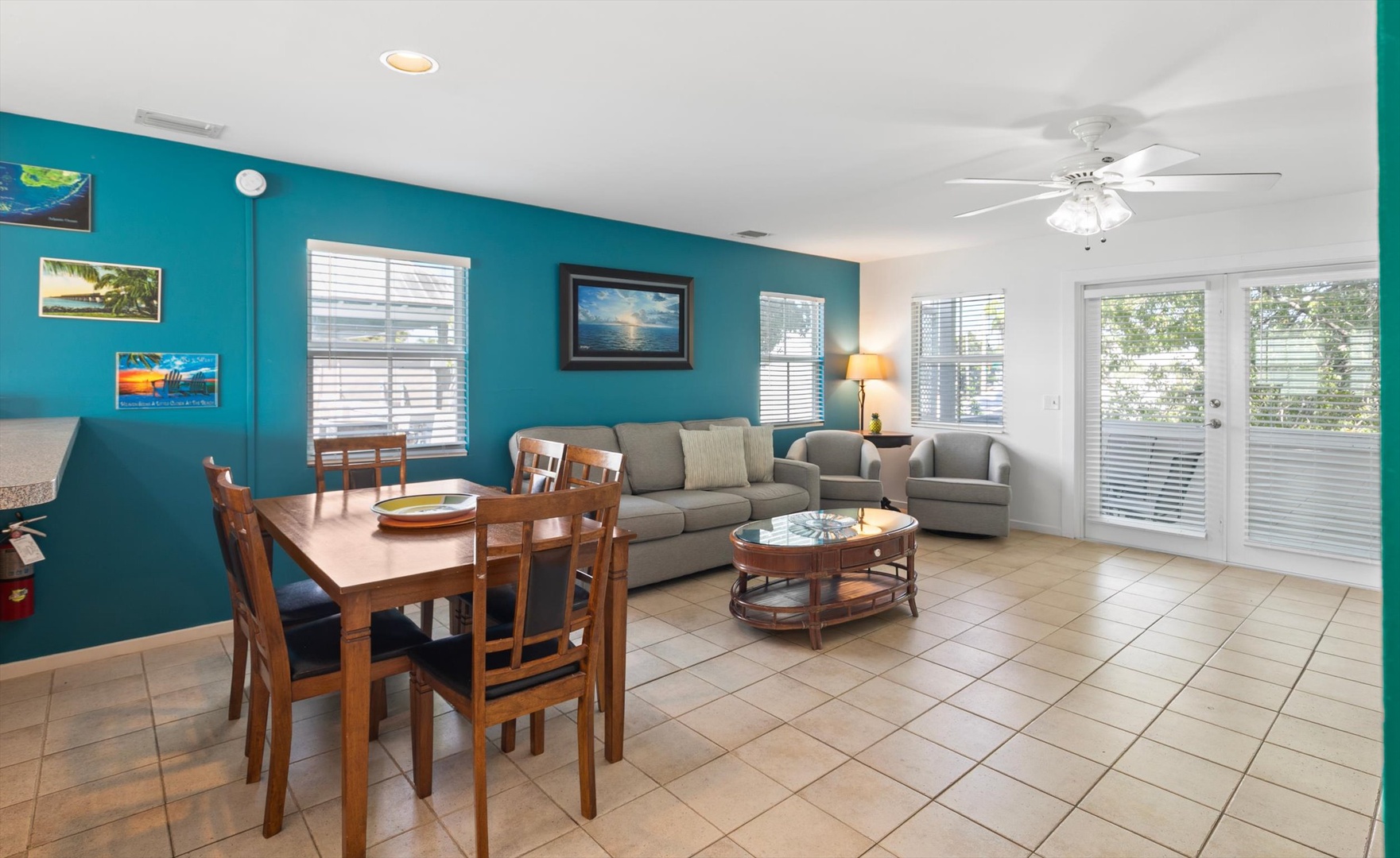 Step into your bright, open living and dining space where coastal-blue walls and abundant natural light create a refreshing, airy feel. Gather around the dining table, relax on the comfortable sofa, and enjoy the breeze from your private patio.