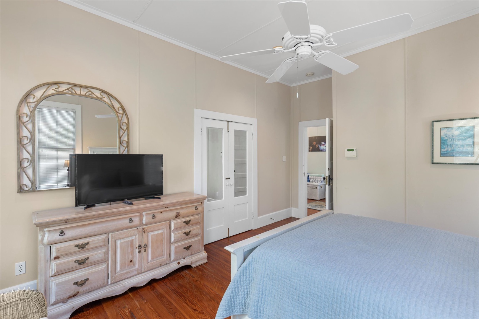 Your serene bedroom features gleaming hardwood floors, a ceiling fan for gentle breezes, and a charming dresser beneath an elegant arched mirror. Natural light fills the space through soft-filtered windows, creating the perfect retreat.